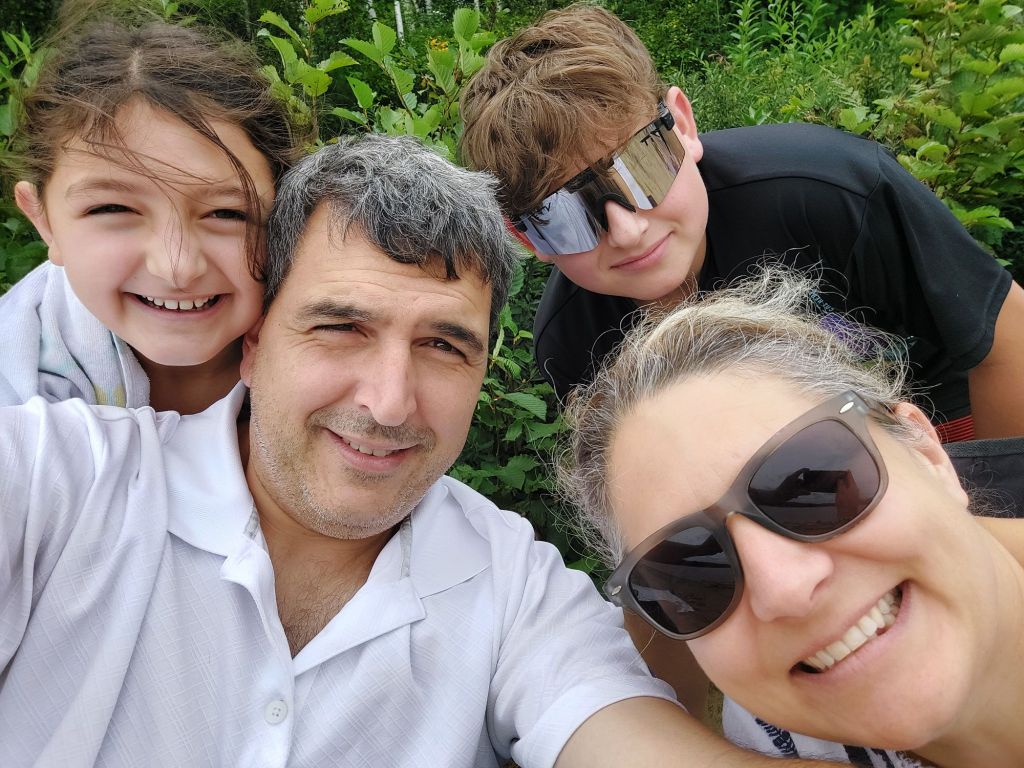 A family selfie featuring four people smiling, surrounded by greenery. The adults are wearing sunglasses, and the children appear happy and cheerful.