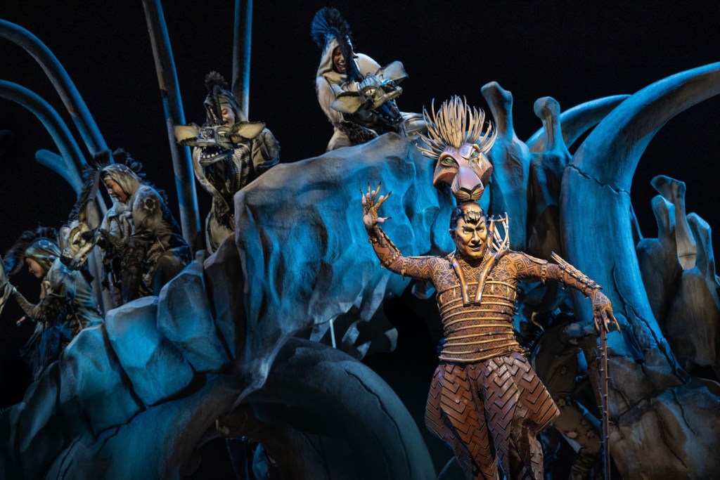 A scene from The Lion King featuring actors in elaborate costumes on stage, with one performer prominently gesturing and others in the background.