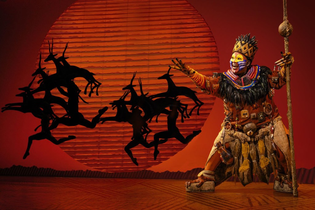 A performer in vibrant tribal costume, holding a staff, stands in front of a large red sunset backdrop while interacting with shadowy animal figures in a theatrical scene from The Lion King.