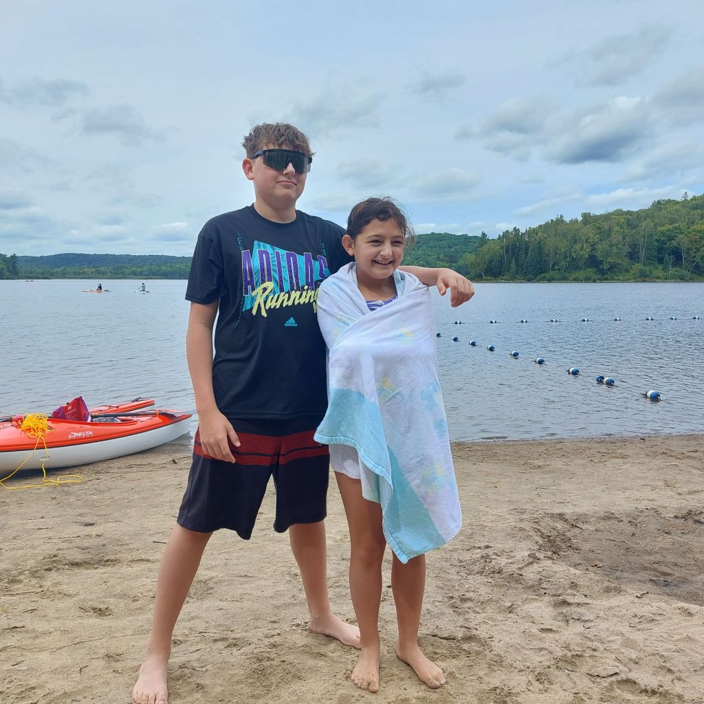 Two children posing at a beach, one wearing sunglasses and the other wrapped in a towel, with a lake and trees in the background.